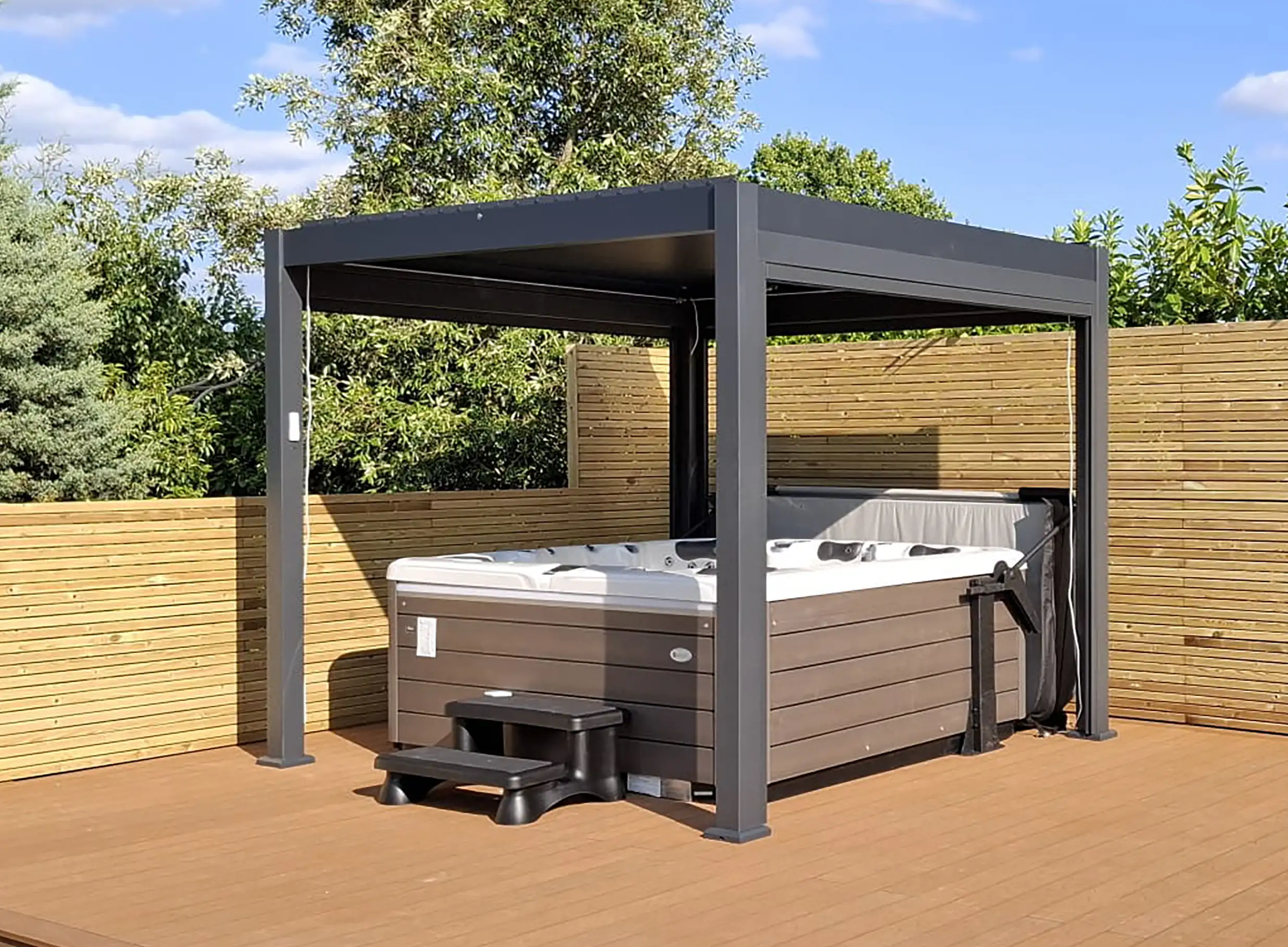 American Whirlpool 881 sat on wooden decking with a grey Maze Living pergola over it for shelter. Hot tub with mocha colour cabinet and white shell. Black steps and a cover lifter.