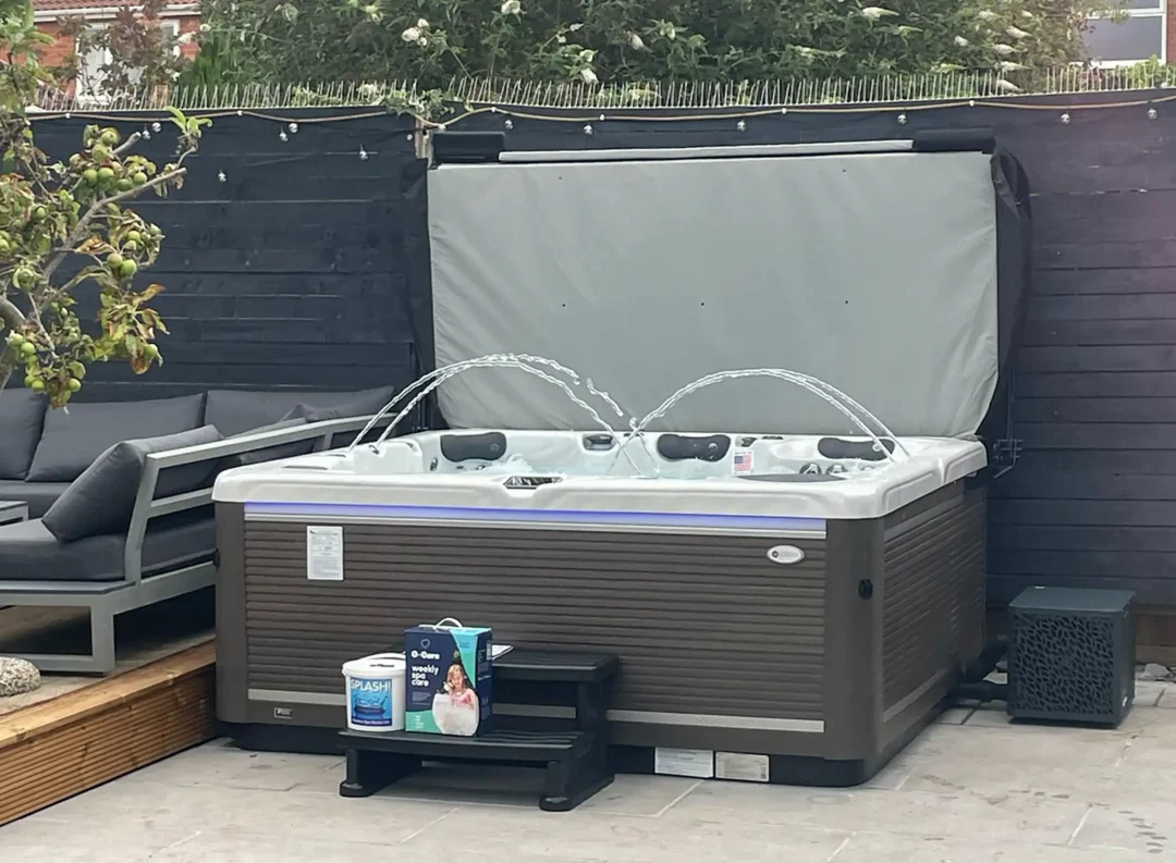 American Whirlpool 881 with a white silver marble shell and charcoal grey cabinet say on patio with black steps and a heat pump fitted. A gas assisted cover lifter fitted for easy opening. 4 Waterfalls arcing over the water and purple LED lights shining