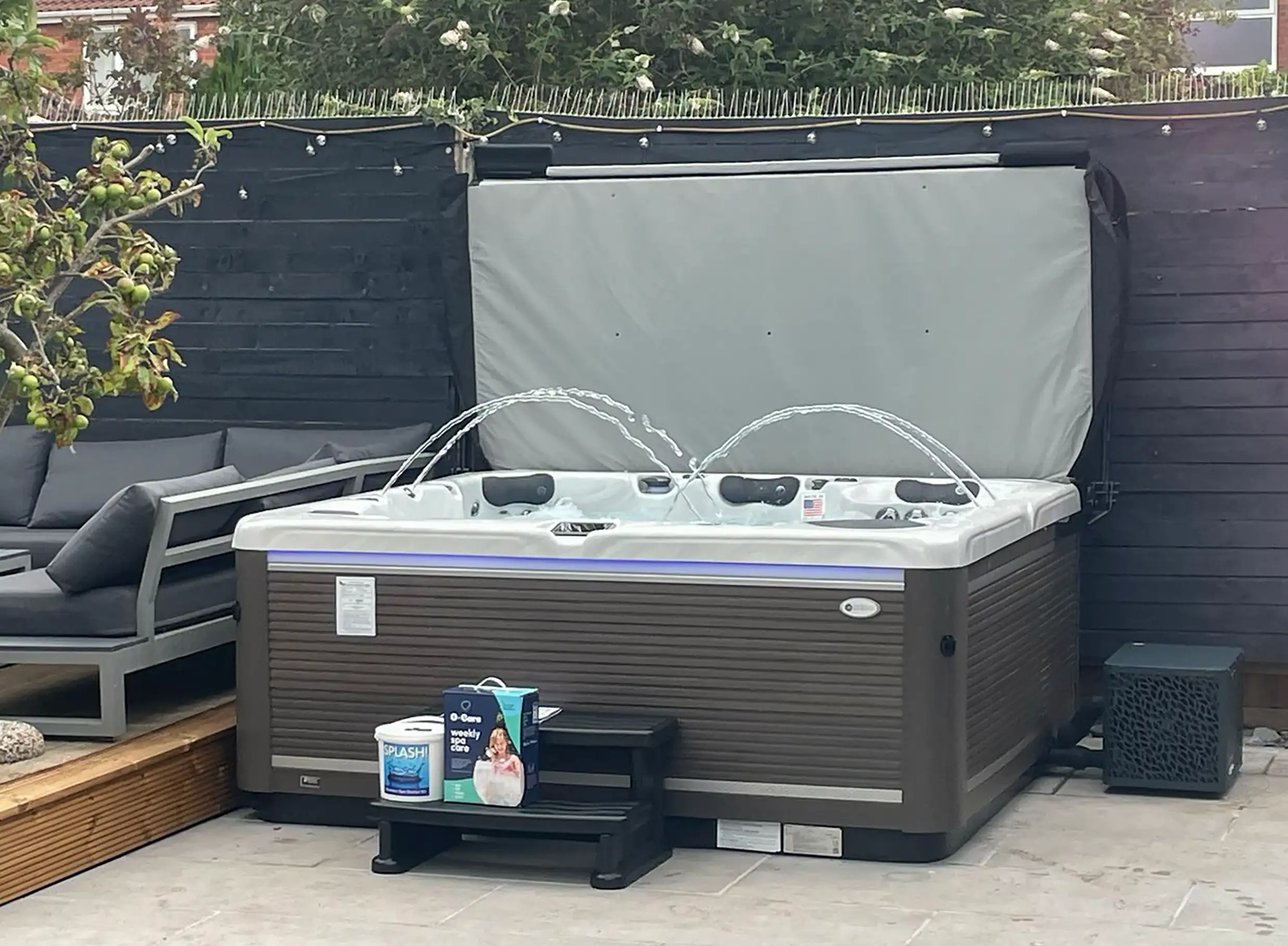 American Whirlpool 881 with a white silver marble shell and charcoal grey cabinet say on patio with black steps and a heat pump fitted. A gas assisted cover lifter fitted for easy opening. 4 Waterfalls arcing over the water and purple LED lights shining