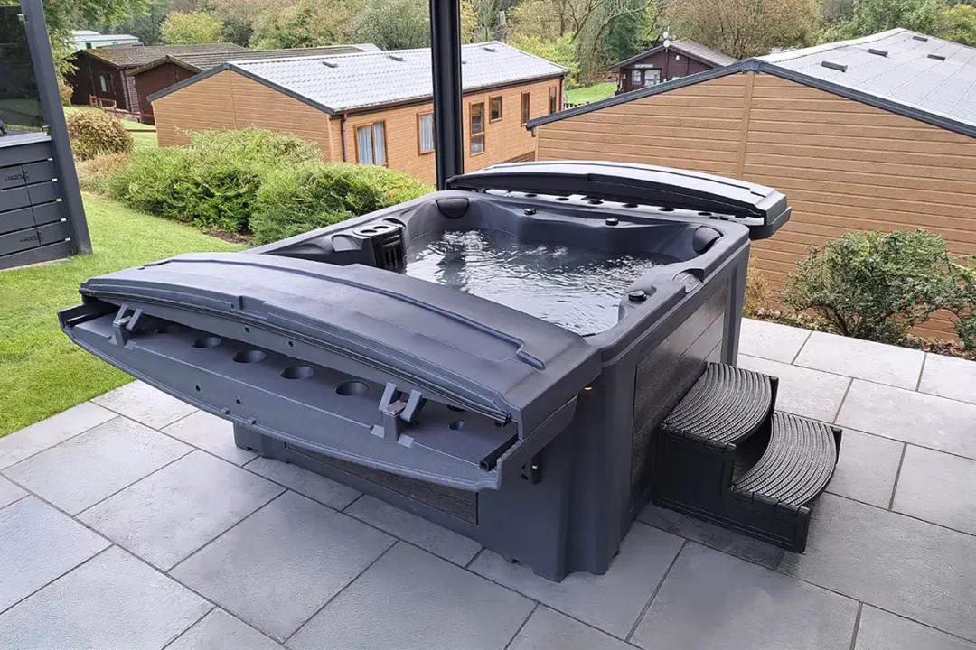 Tuff Spas rotomolded hot tub with tough lid open, steps fitted, all sat on a grey stone patio overlooking holiday lodges